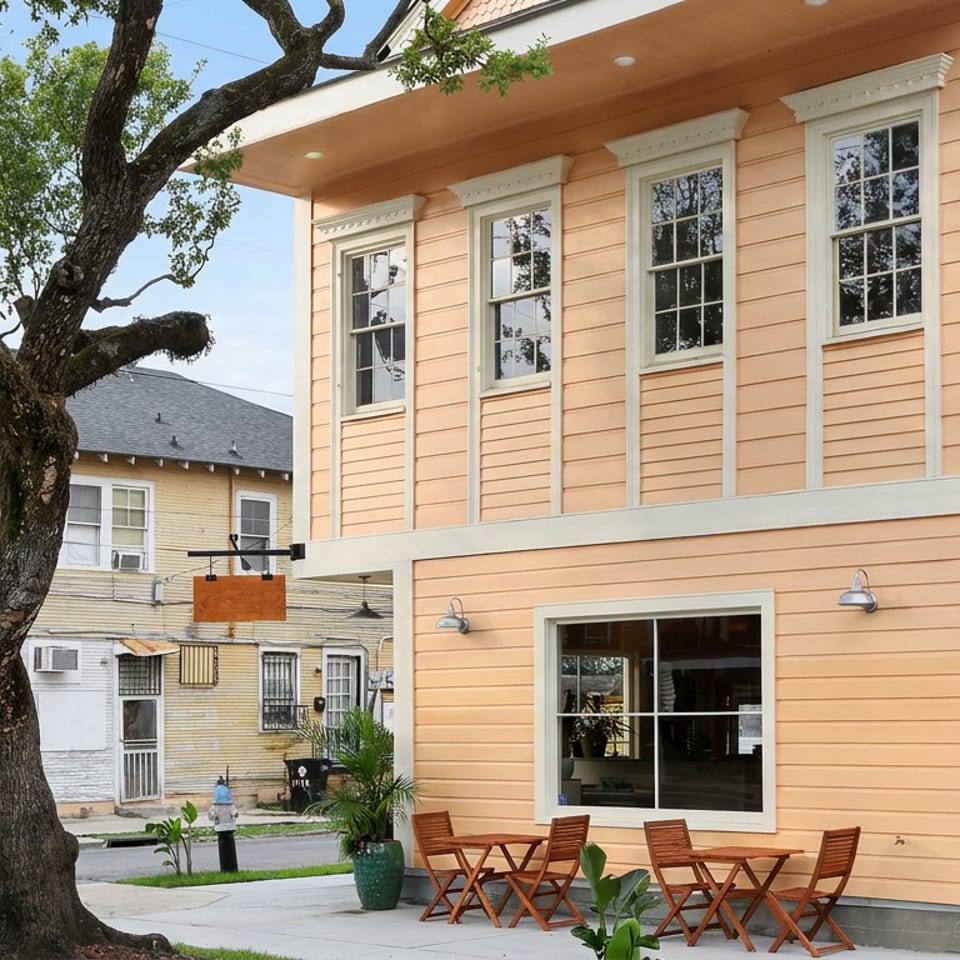 Peach-colored restaurant building with white-trimmed windows, outdoor wooden tables, chairs, and a large shaded tree.