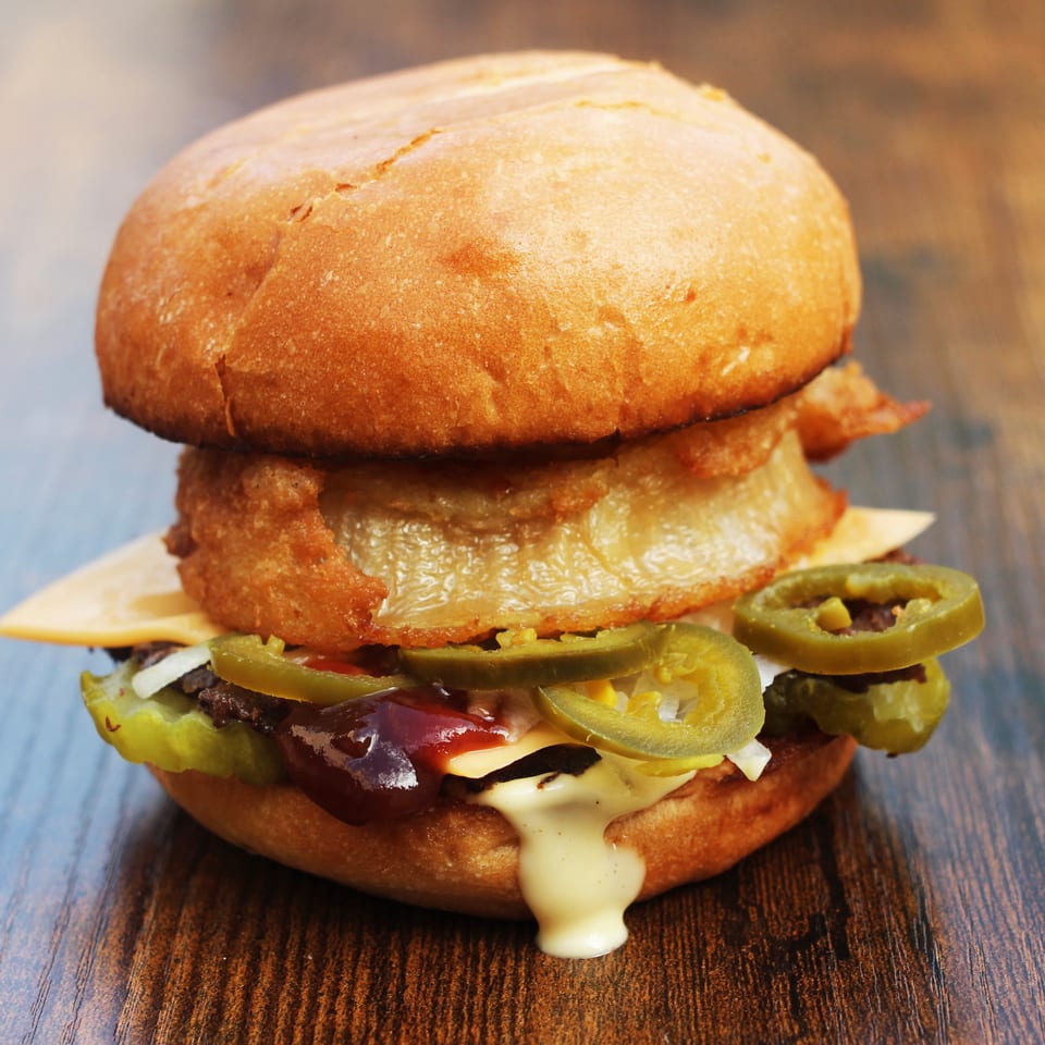 Loaded cheeseburger with a fried onion ring, jalapeños, pickles, barbecue sauce, and melted cheese on a wooden table.