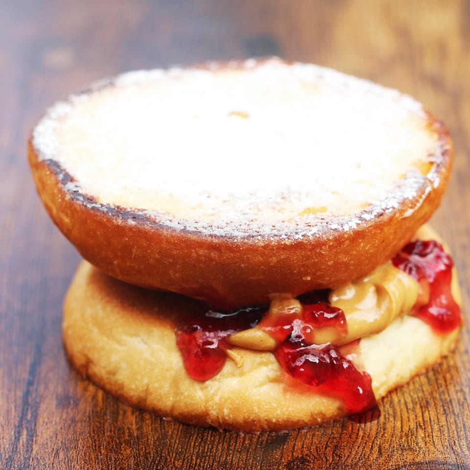 Powdered sugar-topped pastry sandwich filled with creamy spread and red berry jam on a wooden table.