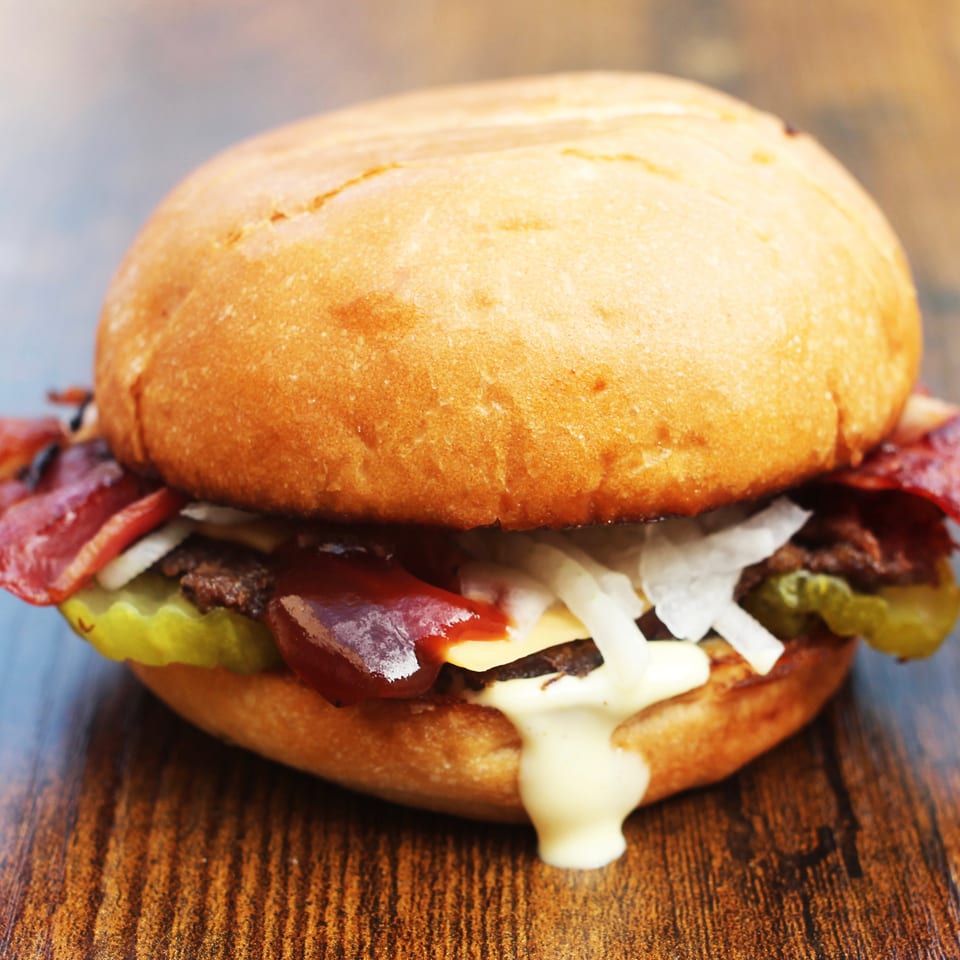 Close-up cheeseburger with bacon, pickles, onions, ketchup, and creamy sauce on a wooden table.
