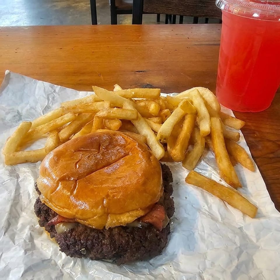 Cheeseburger with a glossy bun, fries, and a red drink on a wooden restaurant table.